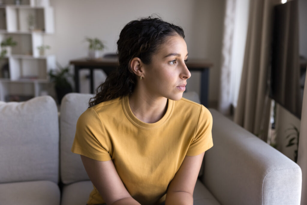 Serious thoughtful 20s Latin woman looking at window away in deep thoughts, sitting on sofa, thinking on problem solving at home.