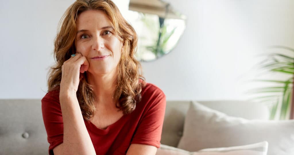 Portrait of a mature woman smiling contently while sitting with a hand on her cheek on a sofa in her sunny home