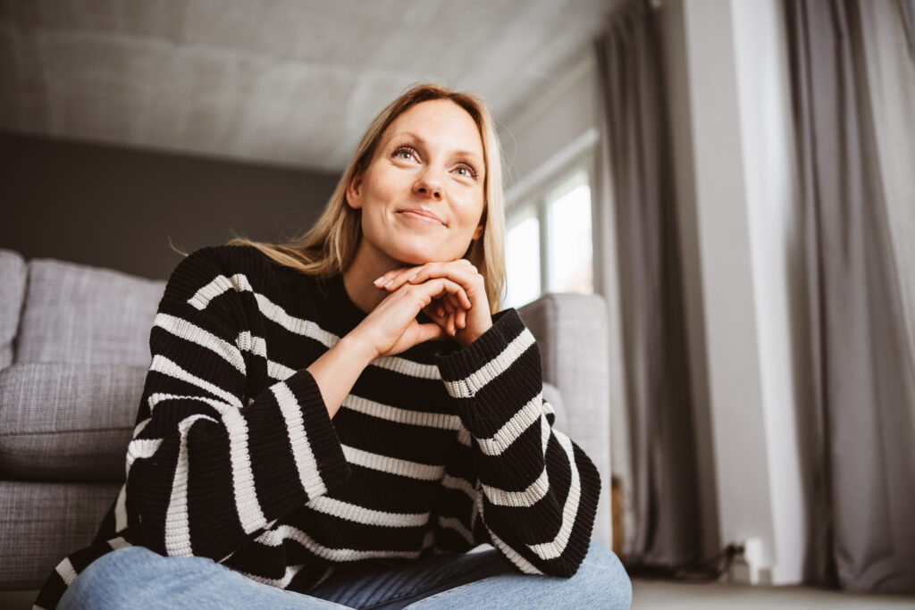Woman in her 30s Sitting on the Floor in Front of Sofa and Smiling at Something Off-Camera; anxiety recovery stories