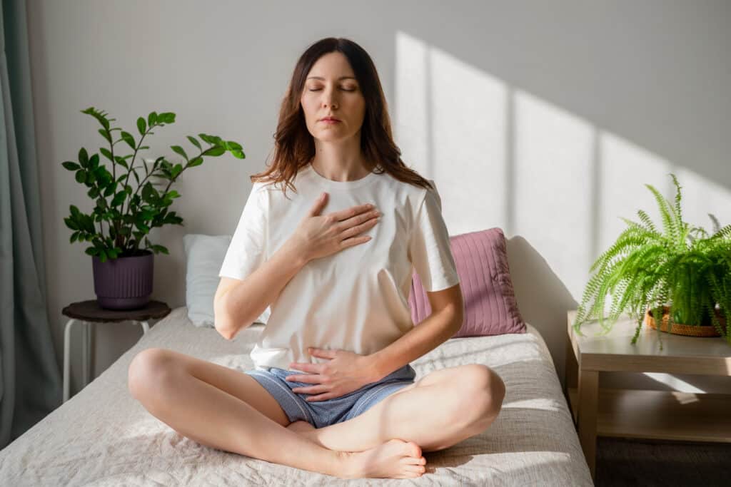 young woman sitting on bed with hand on belly and chest practicing how to stop a panic attack fast