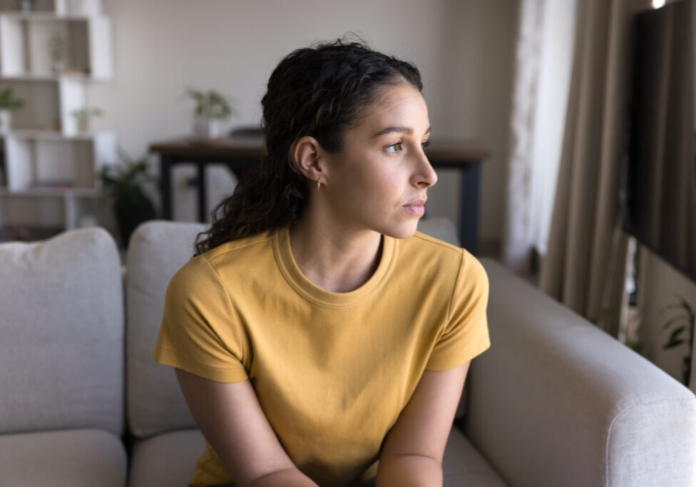 Serious thoughtful 20s Latin woman looking at window away in deep thoughts, sitting on sofa, thinking on problem solving at home.