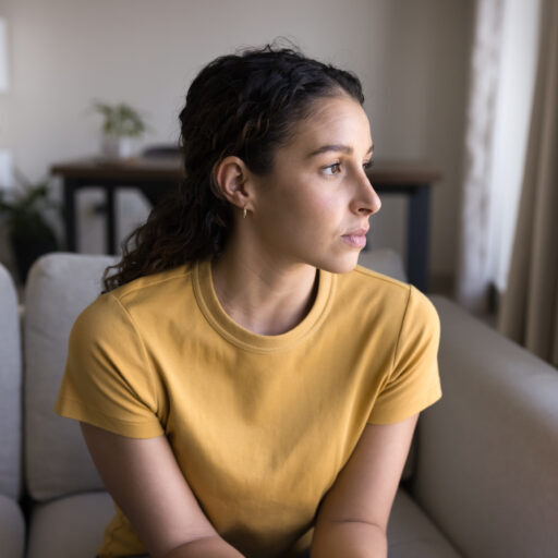 Serious thoughtful 20s Latin woman looking at window away in deep thoughts, sitting on sofa, thinking on problem solving at home.