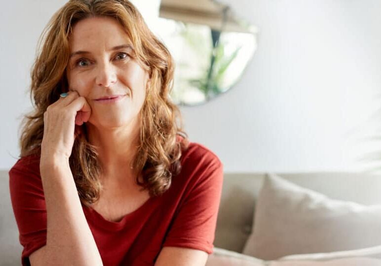 Portrait of a mature woman smiling contently while sitting with a hand on her cheek on a sofa in her sunny home