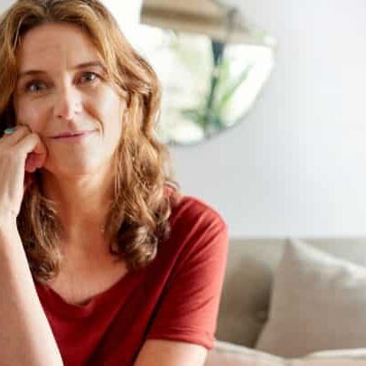 Portrait of a mature woman smiling contently while sitting with a hand on her cheek on a sofa in her sunny home