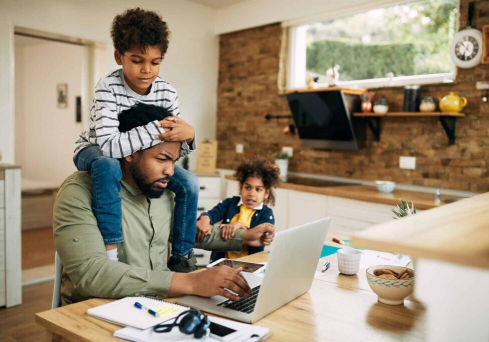Distraught African American father using laptop and working at home while being distracted by his small children.