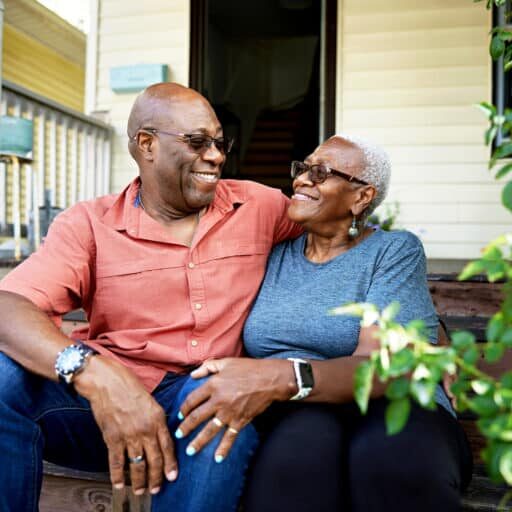 Outdoor portrait of man and woman in casual summertime clothing and sunglasses, sitting with arms around each other and smiling face to face.