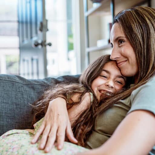 An elementary school age girl sits on her affectionate mother's lap as they cuddle with each other on the living room sofa at home.