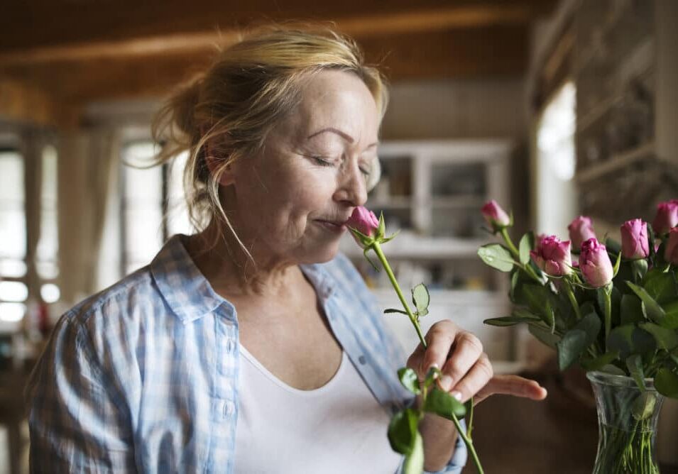 Beautiful senior woman at home in her kitchen smelling a fragrance of pink rose