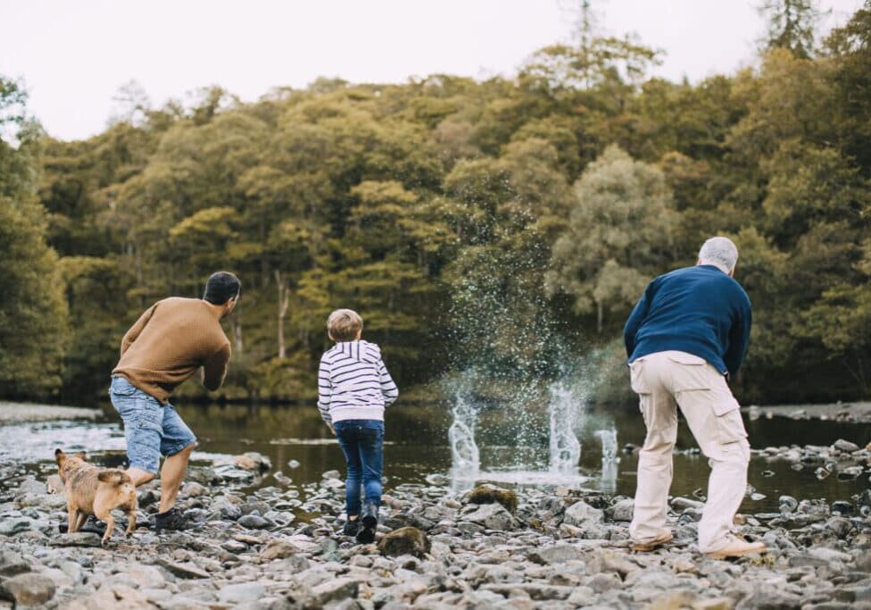 Little boy is skimming pebbles on a lake with his father and grandfather.
