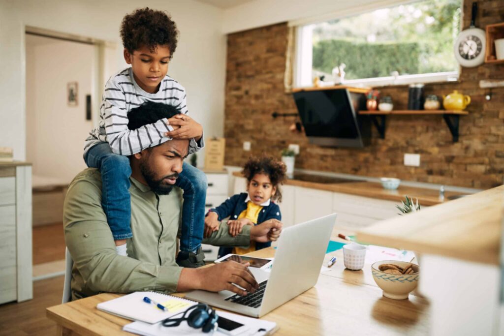 Distraught African American father using laptop and working at home while being distracted by his small children.