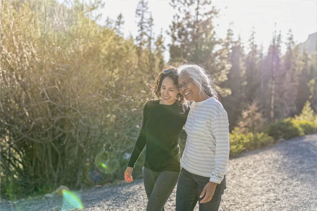 A beautiful mixed race young adult woman embraces her vibrant retirement age mother. The mother and daughter are enjoying a relaxing walk in nature on a beautiful, sunny day. In the background is a mountainous evergreen forest bathed in sunlight.