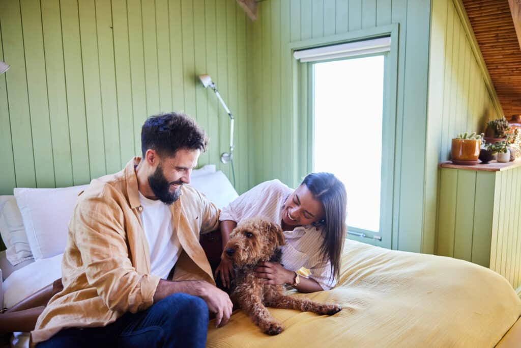 Young couple laughing while petting their dog while relaxing together in their bedroom at home