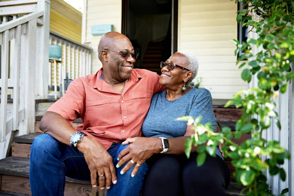 Outdoor portrait of man and woman in casual summertime clothing and sunglasses, sitting with arms around each other and smiling face to face.