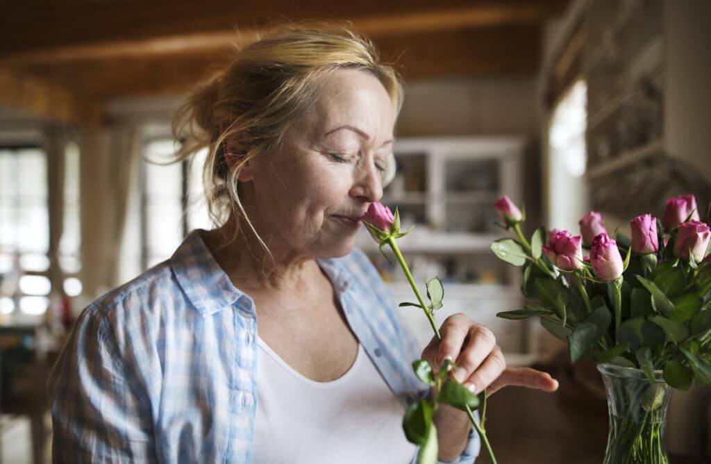 Beautiful senior woman at home in her kitchen smelling a fragrance of pink rose